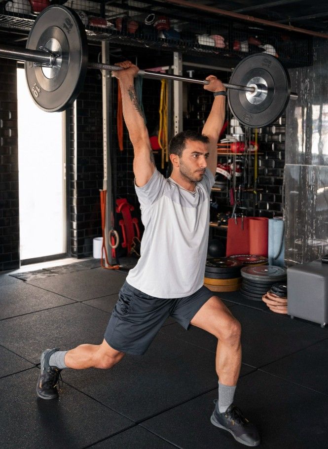 Mann führt einen Ausfallschritt mit einer Langhantel über Kopf aus – funktionelles Krafttraining im Fitnessstudio.