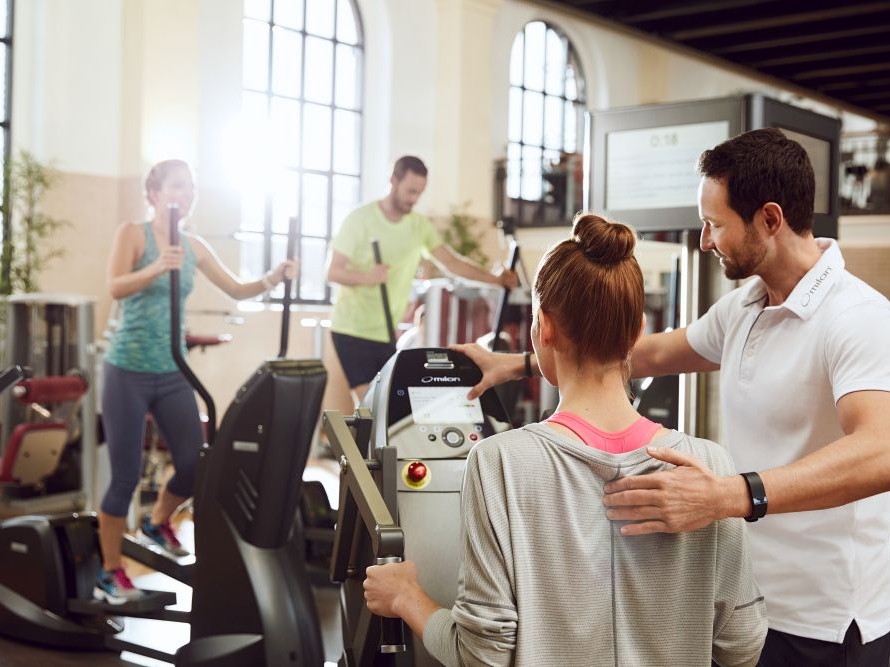 Trainer unterstützt eine Frau an einem Milon-Gerät, während im Hintergrund weitere Personen auf Cardiogeräten trainieren.