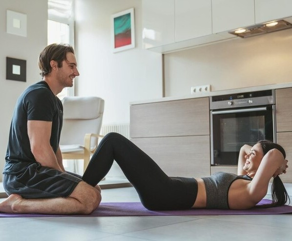 Frau macht Sit-ups auf einer Matte zu Hause, während ein Mann ihre Füße fixiert und sie unterstützt – gemeinsames Training im Wohnzimmer.